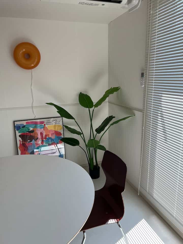 明るい白を基調としたダイニングルーム、丸テーブルと濃い茶色の椅子、壁際の緑の植物が配置されたシンプルな空間