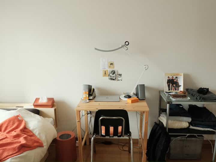 オレンジ色のアクセントがあるシンプルな6畳の寝室兼ホームオフィス、木製の机とオフィスチェアが配置された落ち着いた空間