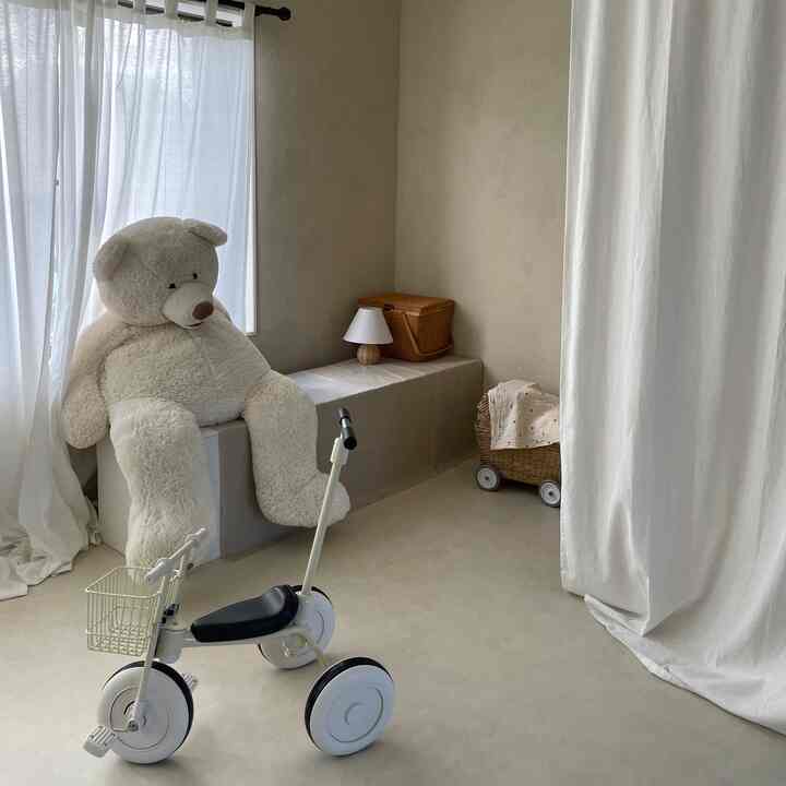 ナチュラルな色調の子供部屋、白いぬいぐるみと三輪車、ラタンの収納バスケットが配置されたシンプルな印象的な空間