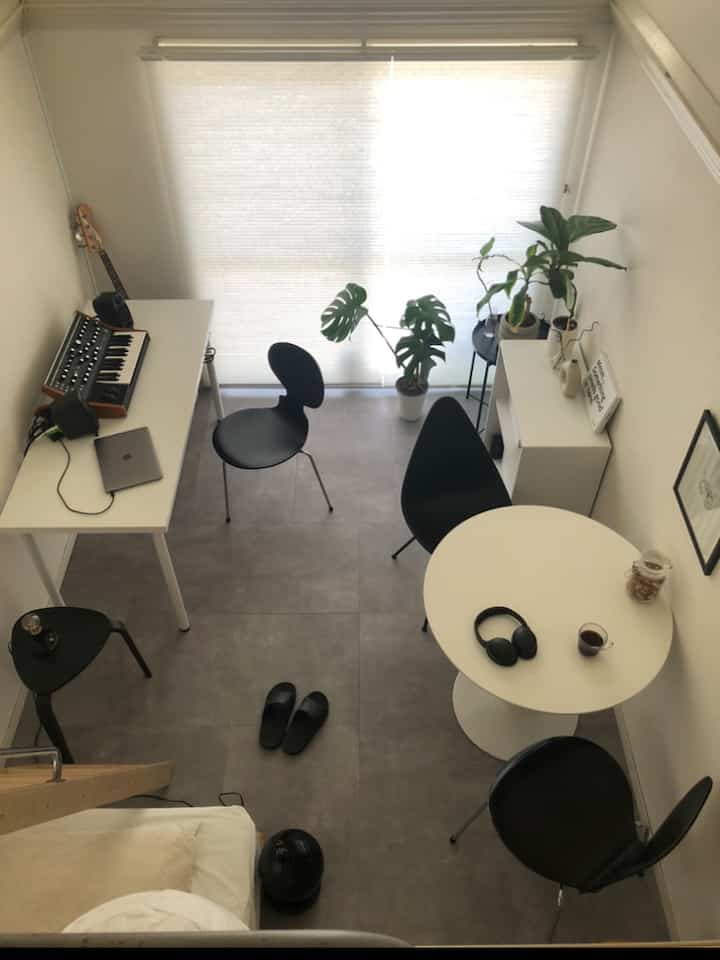 Bright beige and gray-tiled compact single household room featuring a work desk, round dining table with black chairs, and plants near the window creating a simple natural atmosphere