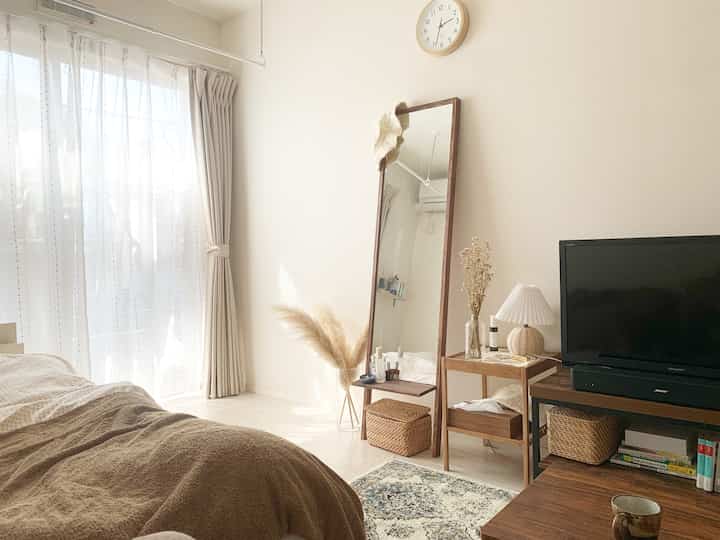 ベージュとブラウンの色調の寝室、長い窓辺にナイトテーブルと全身鏡が配置され、自然な雰囲気のあるインテリア