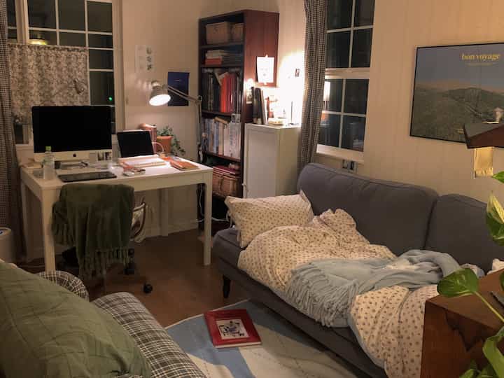 Cozy bedroom-style living room with warm wood tones and gray sofa, featuring a desk, bookshelf, and wall poster