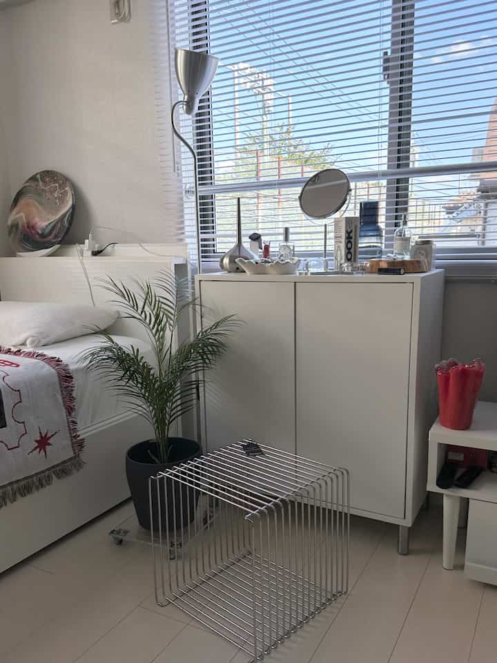 White-toned bedroom featuring a silver wire shelf, floor lamp, and green plant, creating a clean and minimal atmosphere