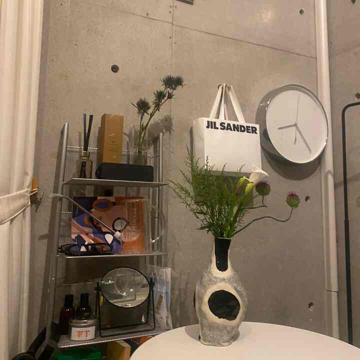 グレーのコンクリート壁と白の丸テーブル、金属の棚と緑の植物が配置されたミニマルなワンルームのインテリア