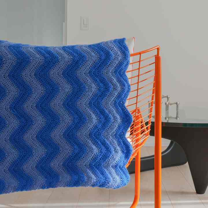 Natural-toned wooden floor living space with white walls, featuring an orange metal armchair draped with a blue crocheted blanket creating a simple atmosphere