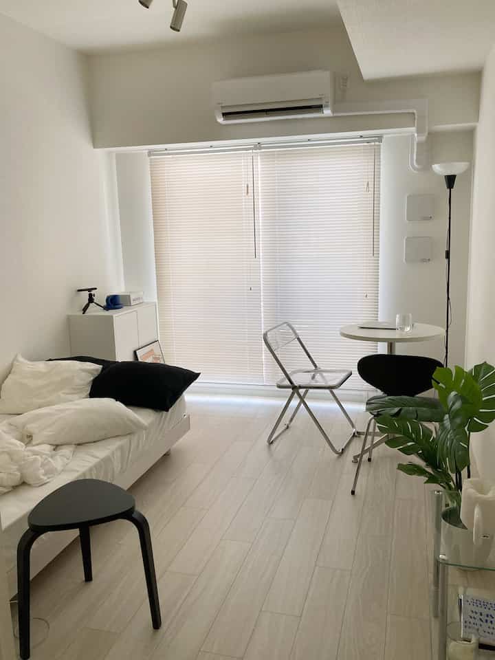 A narrow single bedroom in white and black tones featuring a bed, stool, blinds, and a small table with chairs in a minimal layout