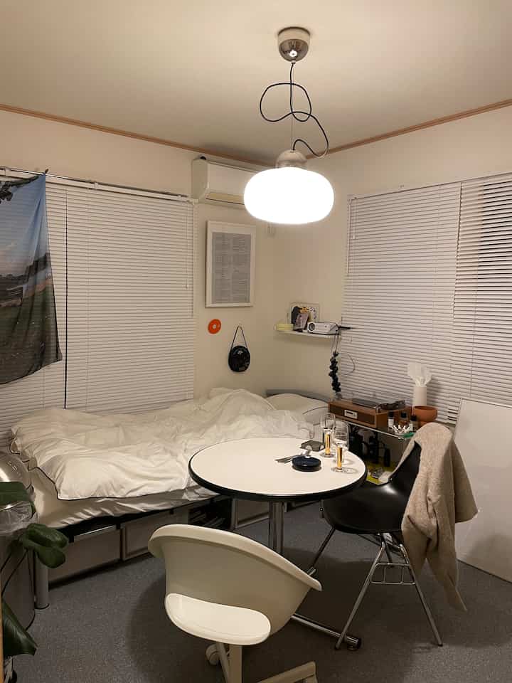 白を基調としたコンパクトなワンルームの寝室、ペンダントライトやダイニングテーブル、椅子が配置された落ち着いた空間