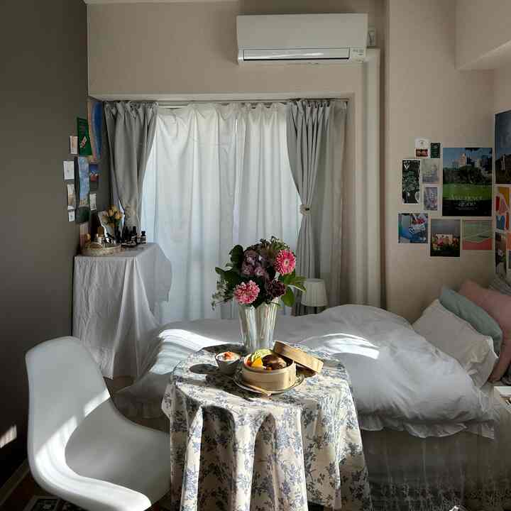 柔らかいベージュとホワイトのトーンの小さな寝室、白い椅子と花の飾られた丸テーブルが配置されたシンプルな空間