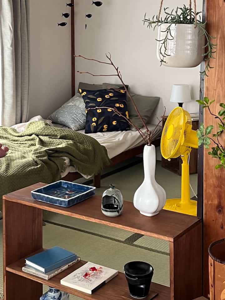 Natural toned Japanese-style studio bedroom featuring a green blanket and yellow standing fan with cozy atmosphere