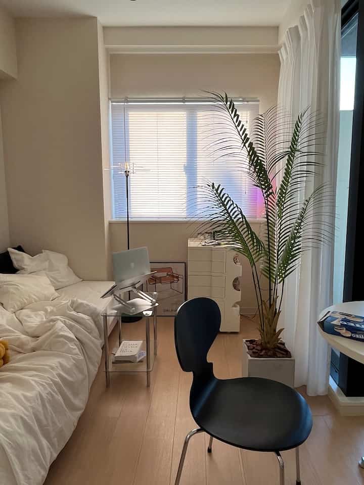 ホワイトとウッドトーンのワンルーム寝室、黒いアームチェアと大きな植物が配置された自然なホームオフィス空間