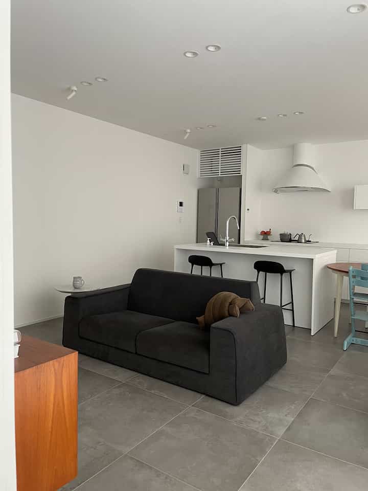 モダンでナチュラルトーンの1LDKリビング・キッチンスペース、黒いソファとカウンタースツール、木製のダイニングテーブルが配置された清潔感のあるインテリア