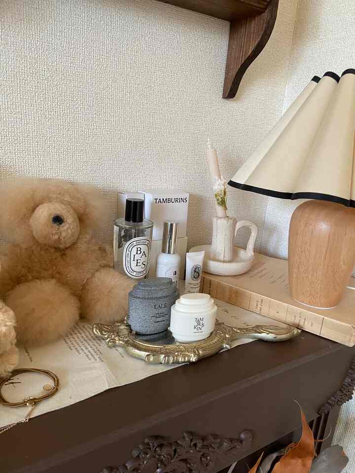 Vintage style display cabinet in beige and brown tones featuring a teddy bear, candle, hand creams, and table lamp with cozy atmosphere