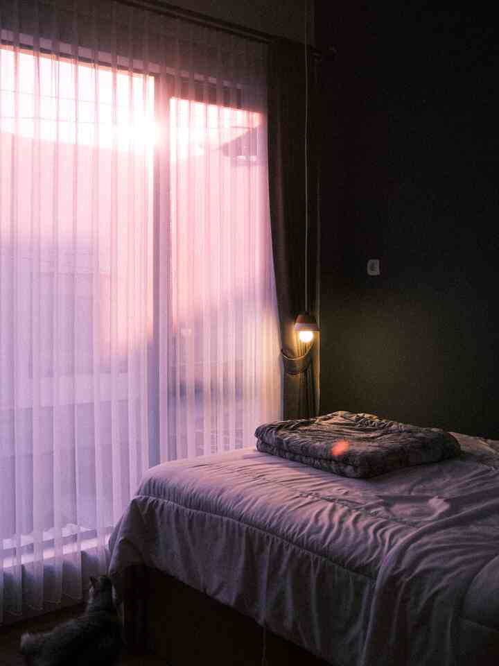 Warm sunset light filters into a bedroom featuring white curtains and bedspread creating a cozy atmosphere