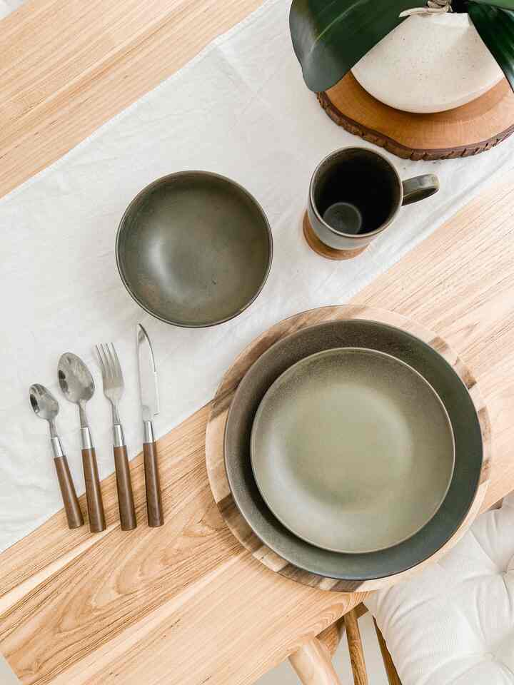 Natural wood tone dining table with white table runner featuring greenish ceramic bowls, plates, mug, and cutlery arranged for a simple warm dining setting
