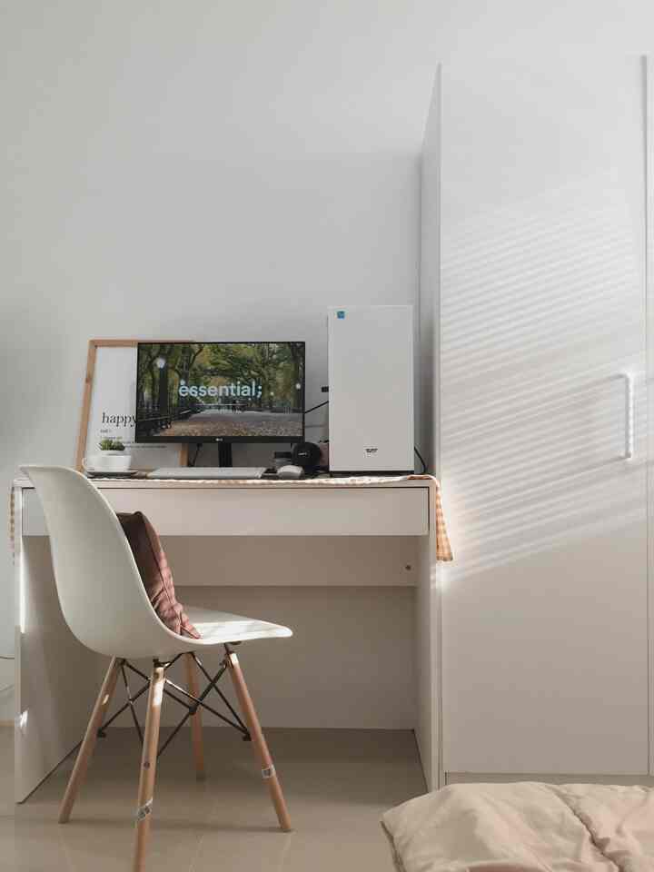 A simple and minimal white bedroom home office featuring a desk with computer, chair, and wardrobe with soft natural light