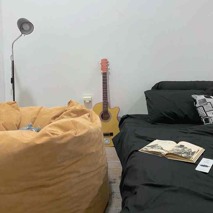 A cozy bedroom featuring a beige bean bag and black bedding, with a guitar and floor lamp in a simple atmosphere