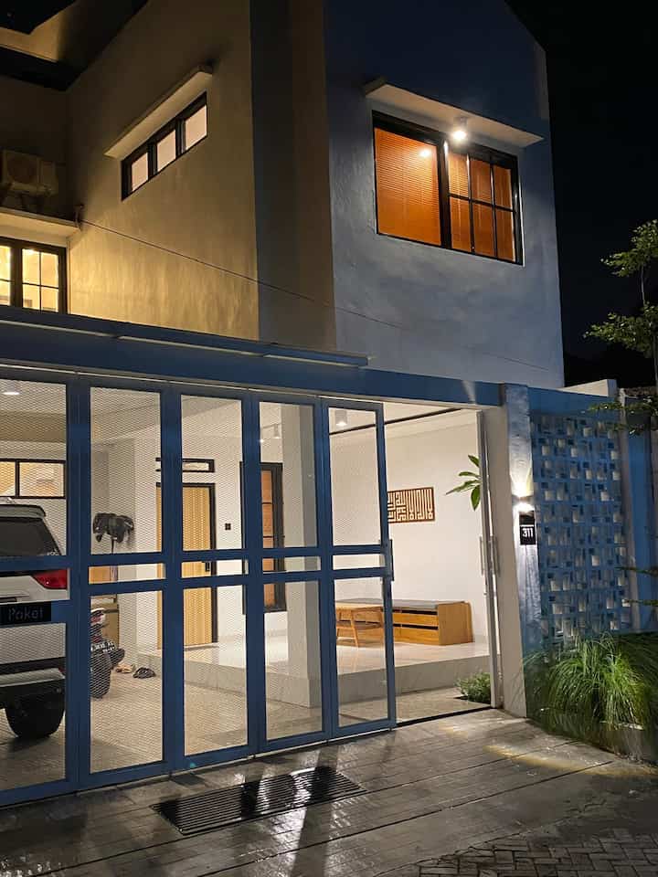 Night-time facade of a modern house with white walls and wood-tone furniture inside, ambient lighting creating warm entrance atmosphere
