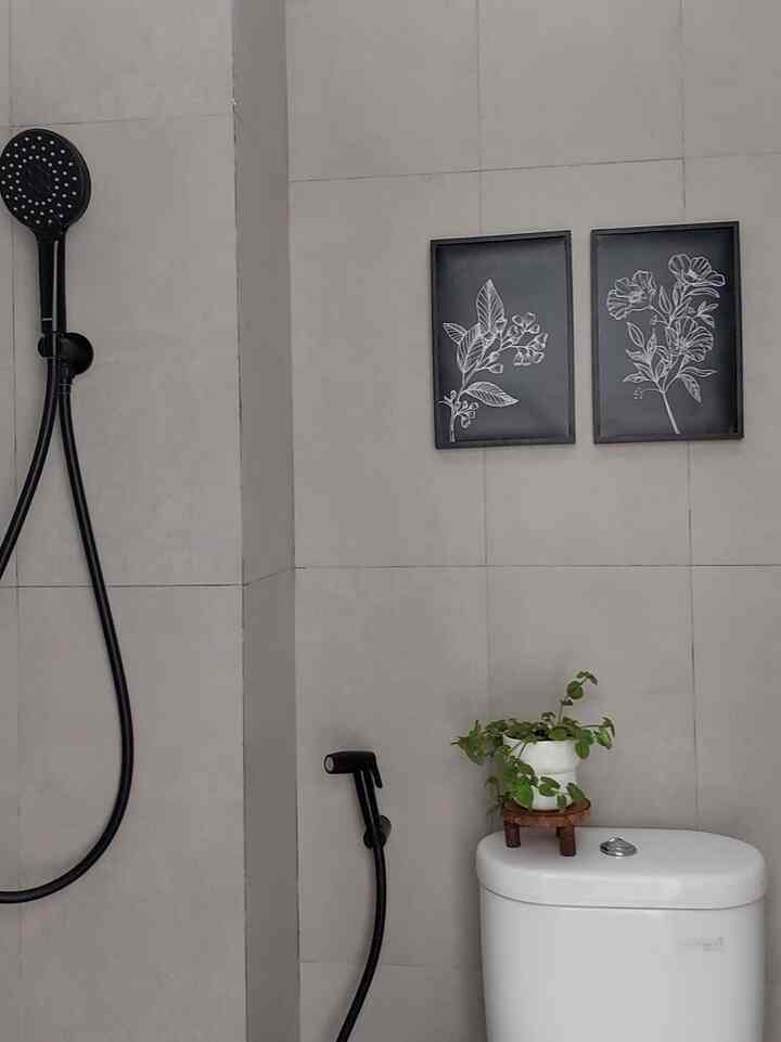 Minimalist small bathroom with light gray tiled walls, white toilet, black shower fixture, potted plant, and framed botanical art