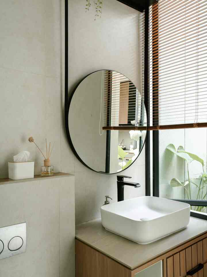 Modern bathroom washbasin area in white and wood tones featuring a black round mirror and diffuser, with a neat and clean interior