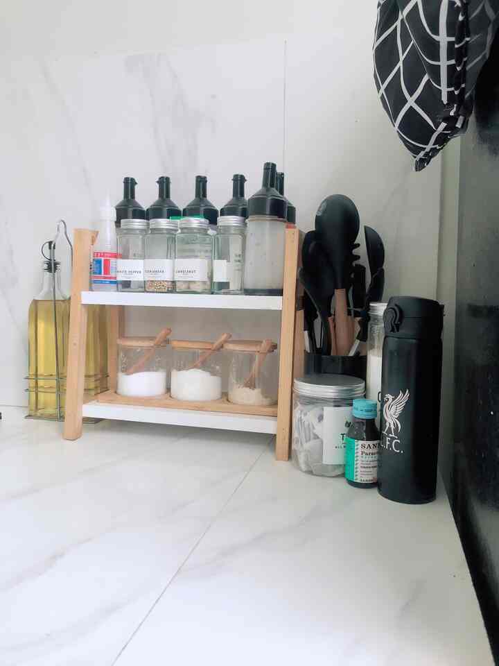 Compact kitchen corner with white flooring and wood-toned shelving, featuring transparent spice jars and black kitchen utensils neatly organized