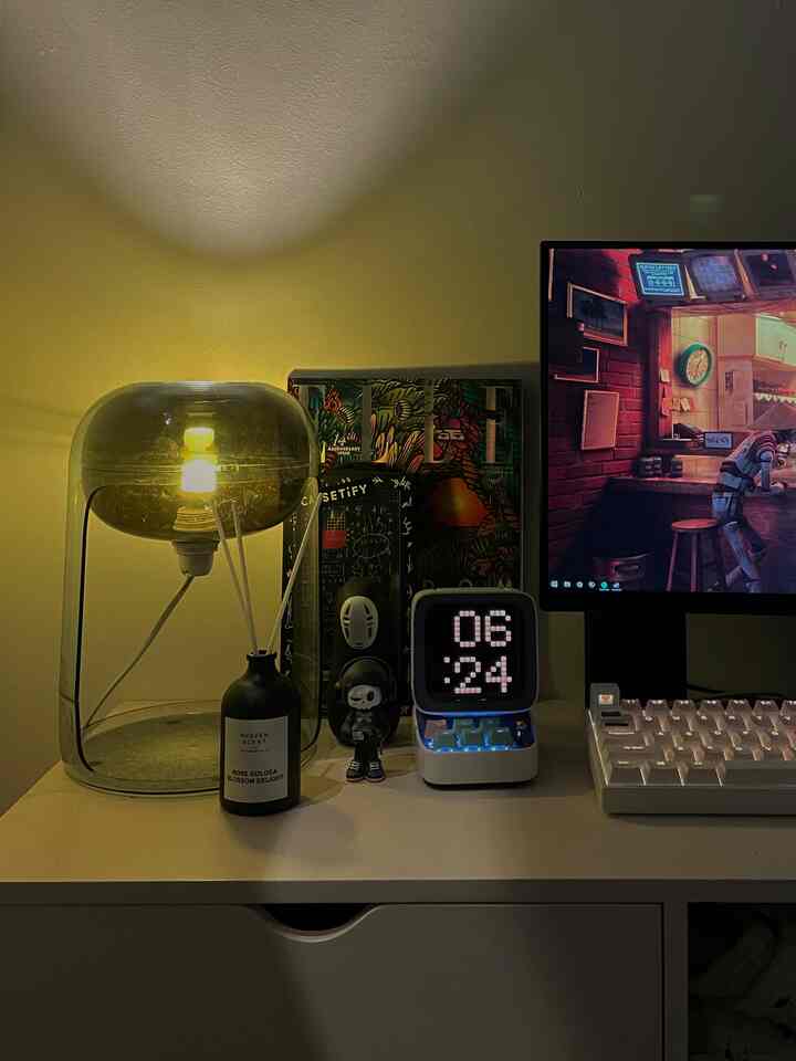 Warm yellow-lit white desk with table lamp, diffuser, digital clock, and gaming keyboard in a home office setup