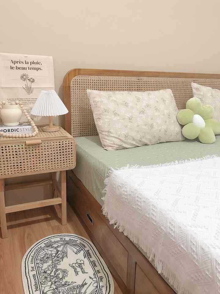 Soft brown and white toned bedroom featuring a rattan headboard bed, nightstand with table lamp, floral pillows and a flower-shaped cushion creating cozy atmosphere