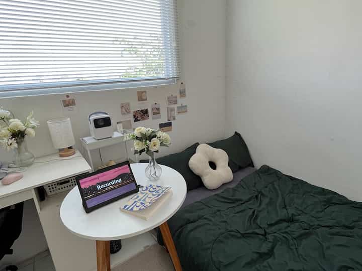 A small bedroom in white and green tones featuring a white desk and round dining table with a simple, minimal atmosphere