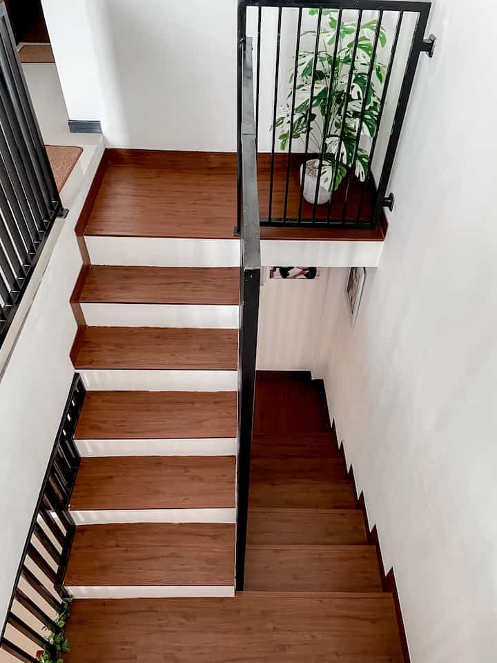 Brown wood-tone staircase with white walls, black metal railing, and an artificial plant creating a clean modern interior space