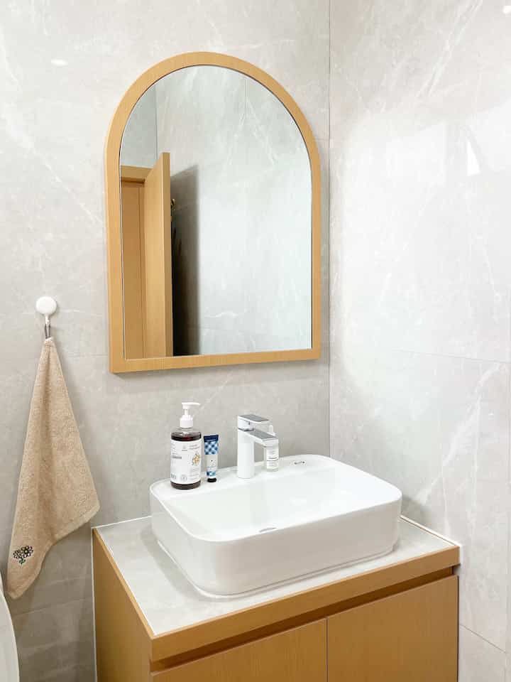 Minimal bathroom featuring light wood tone vanity and arched mirror, white wash basin, and gray tiled walls creating a clean, serene atmosphere