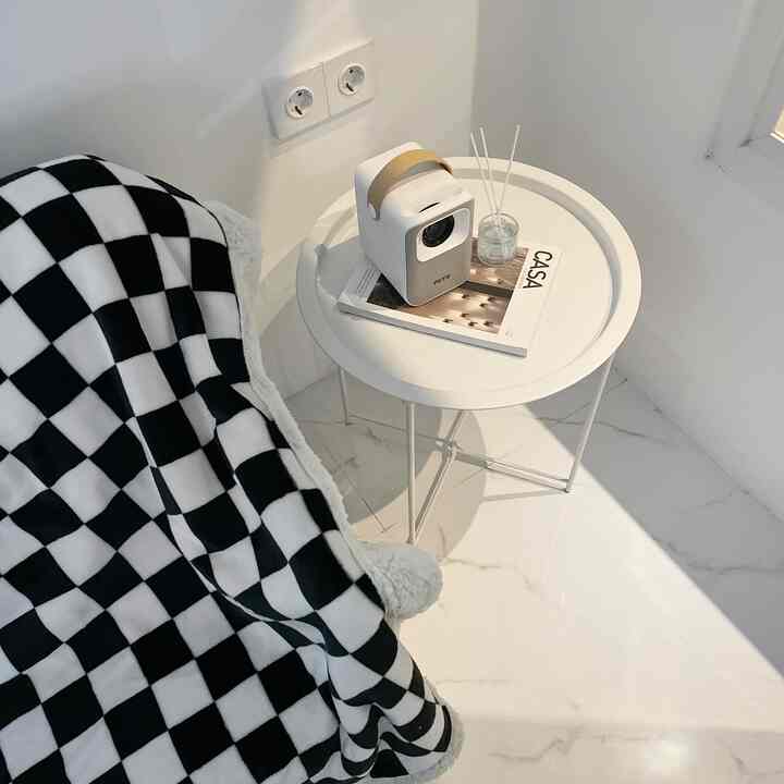 White-toned living room featuring a round white side table with a projector and diffuser, and a black-and-white checkered blanket draped over a sofa, creating a simple space