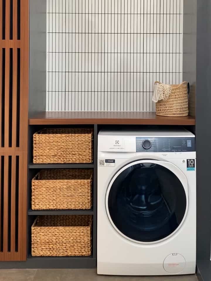 A compact laundry room with white tiled wall, brown wooden countertop, neatly organized woven baskets, and a washing machine