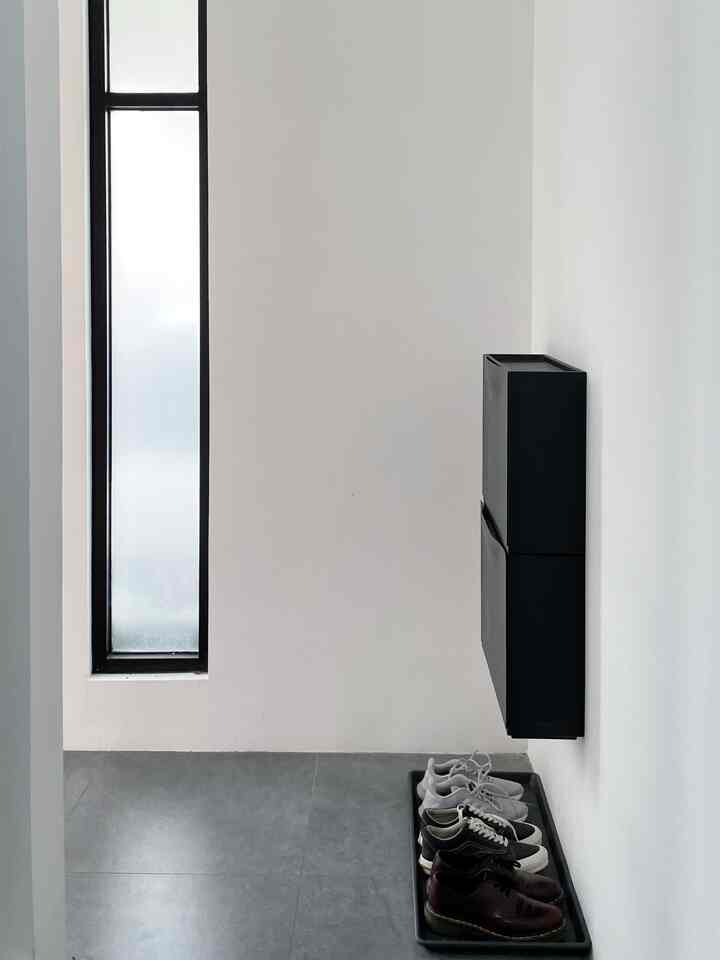 Monotone entrance with white walls and gray floor, black shoe cabinet mounted on wall and neatly arranged shoes on floor tray, creating tidy storage space