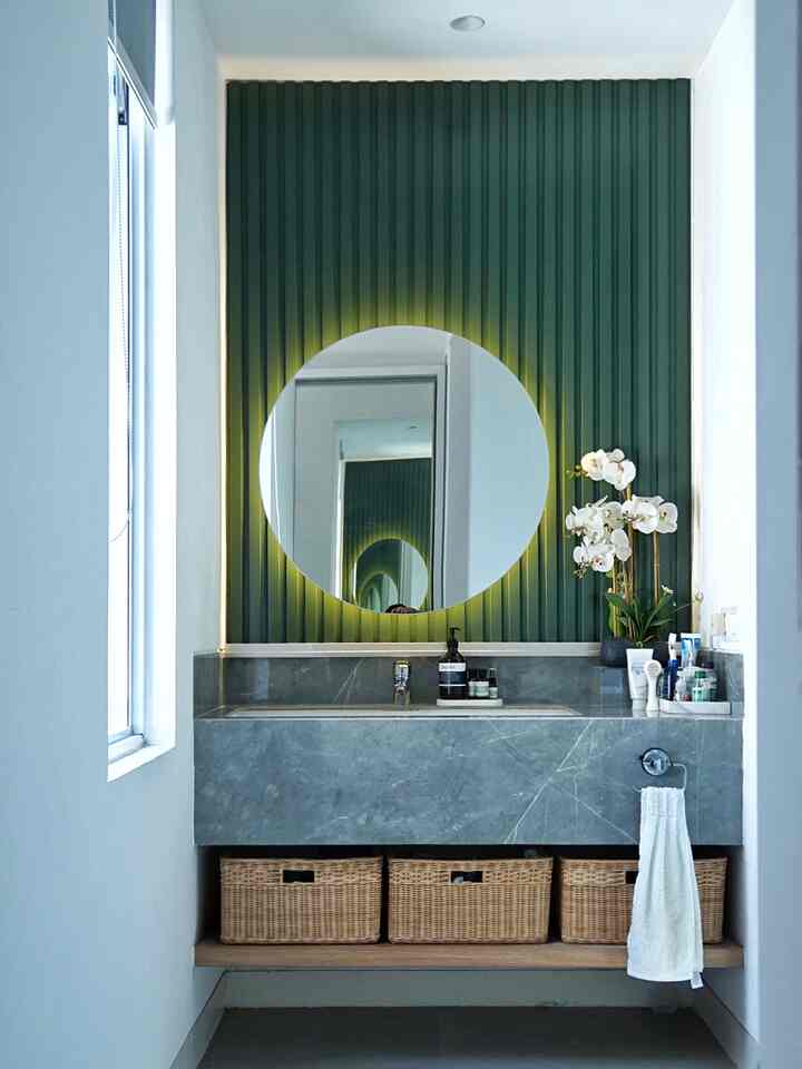 Natural modern bathroom featuring gray marble vanity and green textured wall with round illuminated mirror