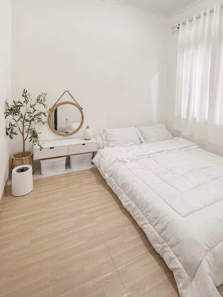 Bright bedroom with white and wood tones featuring bed, round mirror, and plant in minimalist style