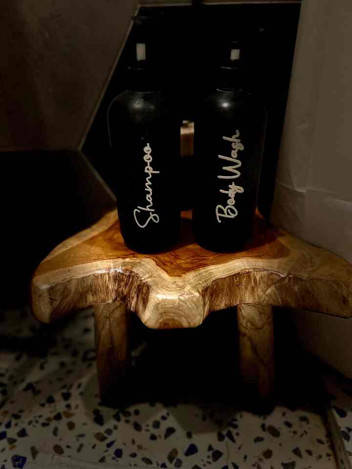 Warm wood tone stool holding black shampoo and body wash bottles in a dimly lit bathroom setting