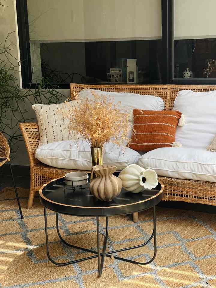 Natural toned terrace featuring rattan sofa, gold vase on black round table, and warm wood-tone carpet creating cozy atmosphere