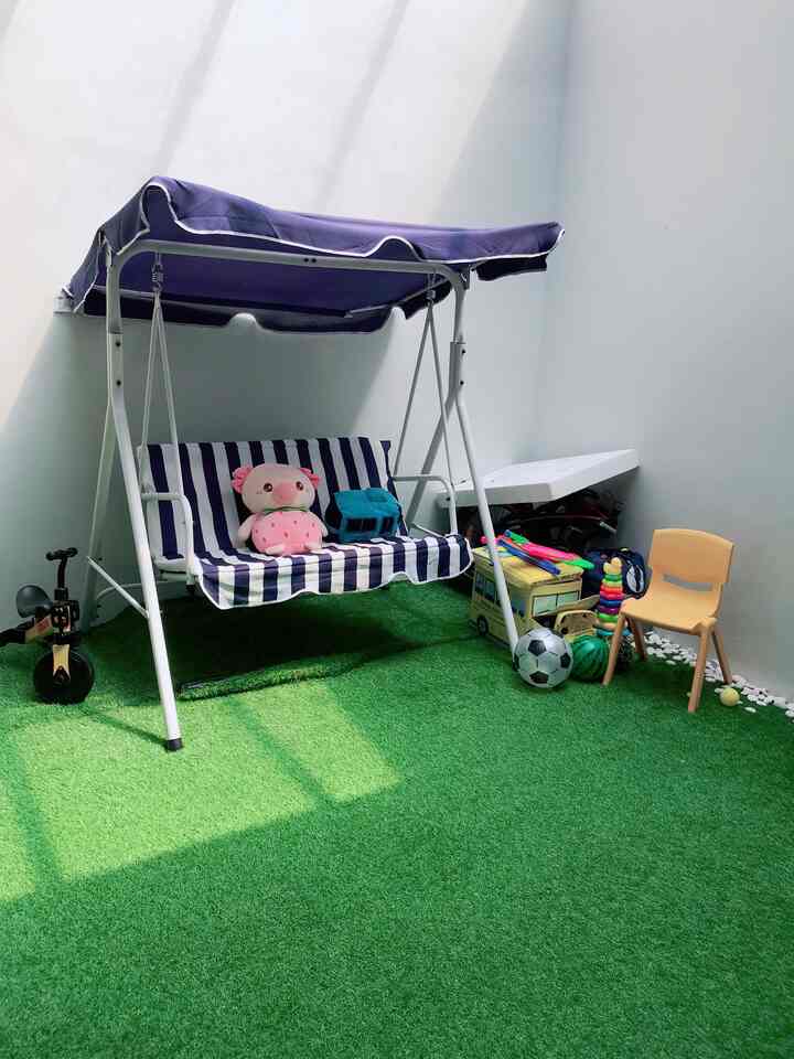 Indoor play area with green synthetic grass carpet and a blue canopy swing seat for three, featuring kids' toys and chair