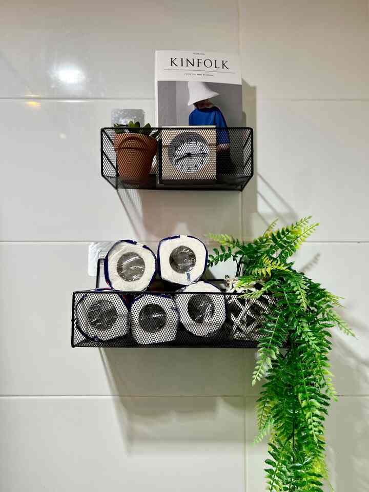 White tiled bathroom wall with black mesh shelves holding plants, clock, and toilet rolls, creating a neat organized space