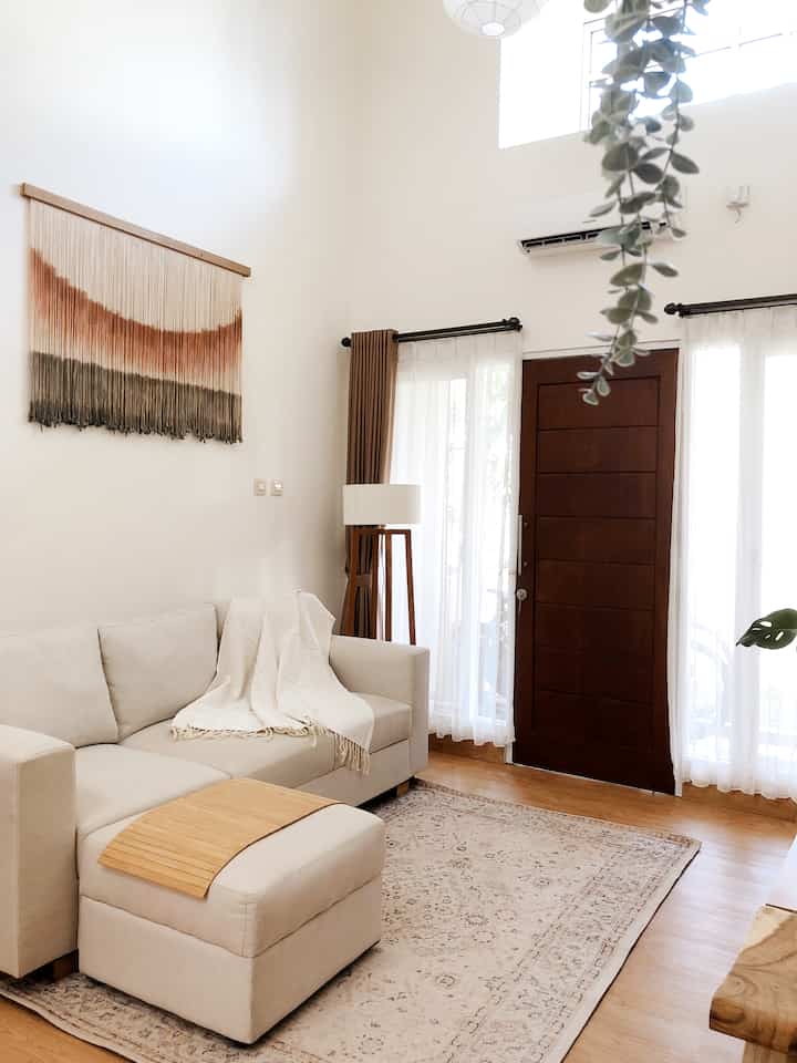 Bright white and brown toned living room featuring beige sofa, carpet, wooden floor, and macrame wall art with a natural cozy atmosphere