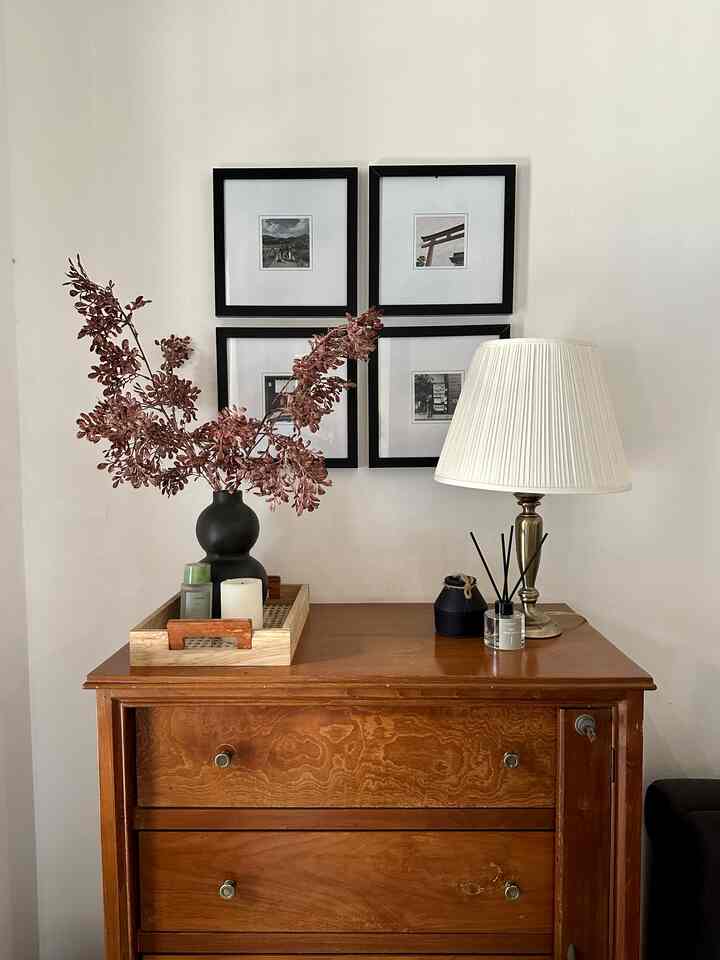 Natural wood tone entrance space featuring a wooden chest, cream table lamp, black vase with dried flowers, and framed pictures creating a cozy atmosphere