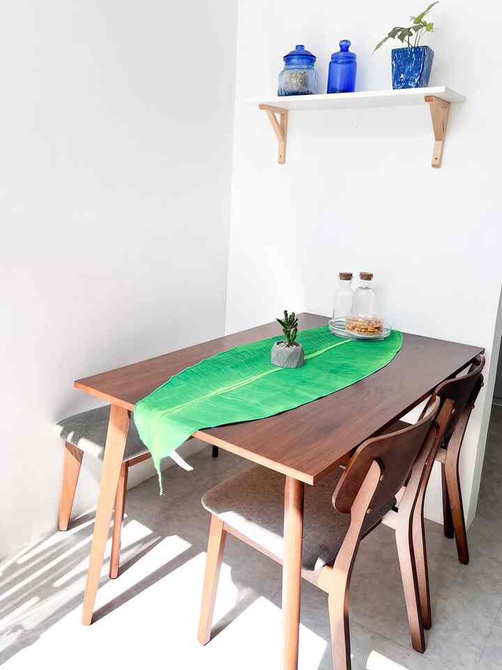 Bright white walls and wood-toned furniture in a 4-person dining room featuring a green leaf-shaped table runner in a minimal natural style