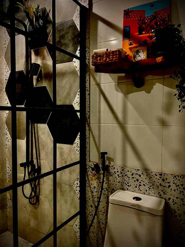 Moody dimly lit modern bathroom with black metal shower frame, hexagonal gray tiles, toilet and wall shelf under warm yellow lighting creating cozy ambiance