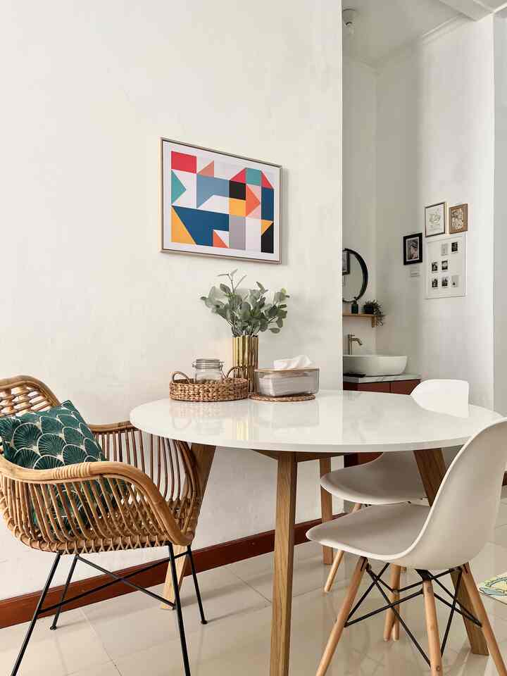 White and natural-toned dining room featuring a round table, rattan chair with cushion, and gold vase accent in a clean setting