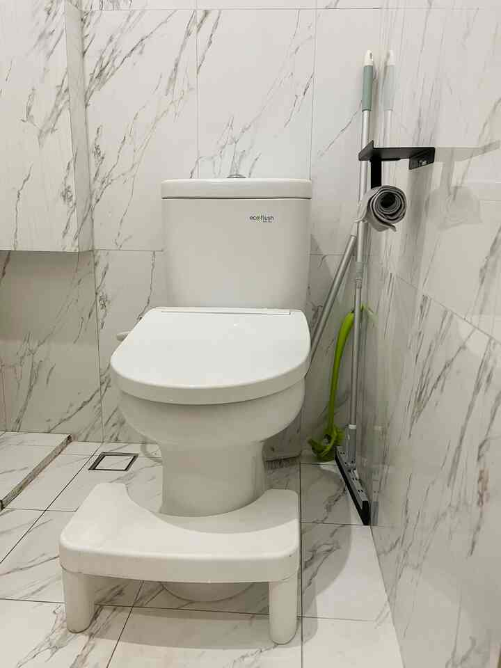 Compact restroom with white and gray marble tiles, featuring a central white toilet and a foot stool positioned in front, creating a clean look
