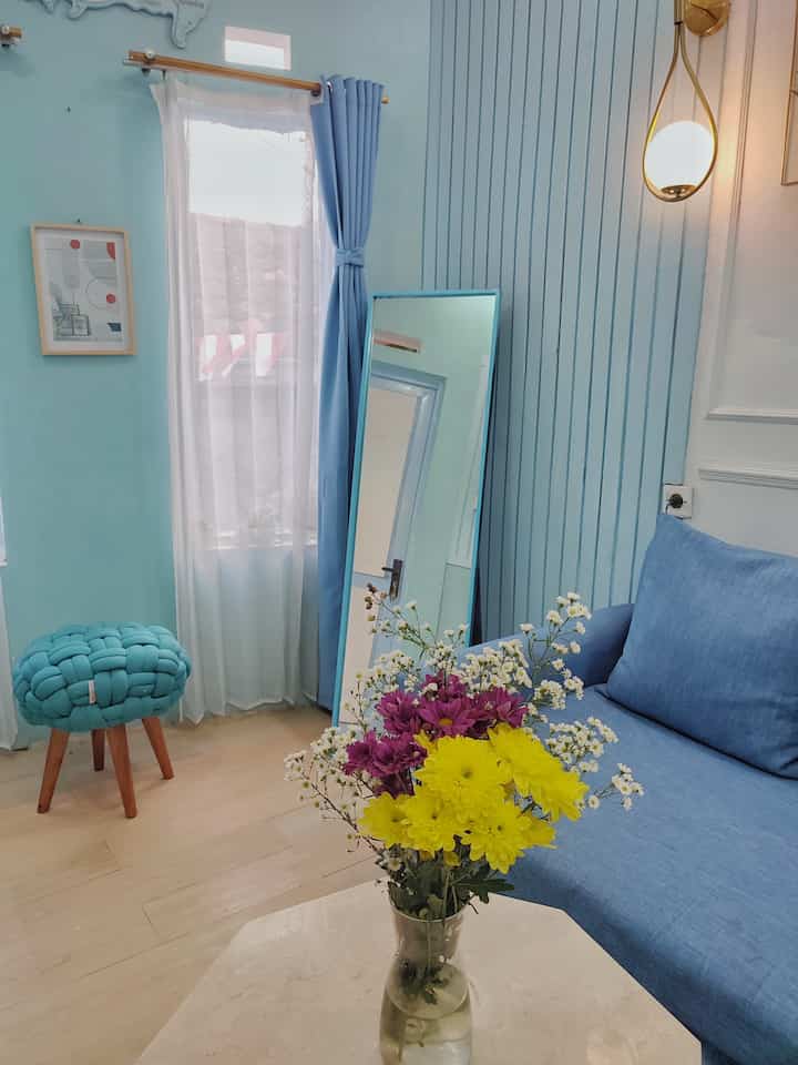 Soft blue-toned small living room featuring a blue sofa, blackout curtains, small stool, and floral arrangement with a simple ambiance