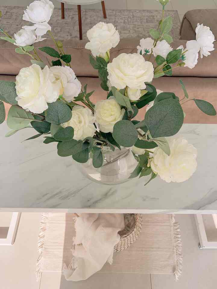 A white and natural toned living room featuring a marble table with white flower decorations and a cozy blanket, creating a warm atmosphere