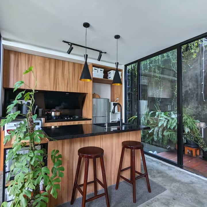 A natural wood and black toned kitchen and home bar featuring a black kitchen island, pendant lights, bar stools, and large glass windows revealing outdoor greenery.
