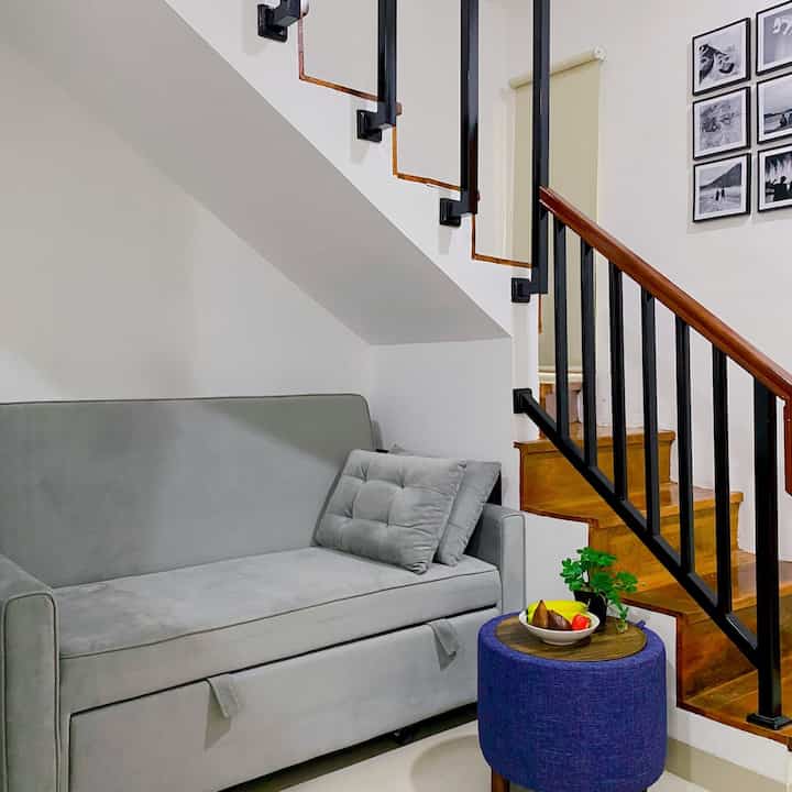Gray sofa bed and blue stool in a compact, simple living room space under wooden stairs with black railing