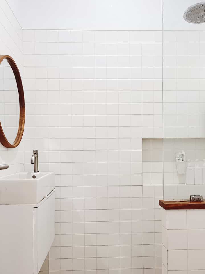 Minimal white and wood tone bathroom featuring shower area, sink, and round wooden-framed mirror with clean atmosphere
