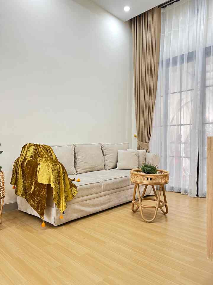 Bright living room with white walls and brown wood-tone flooring, featuring a beige sofa and rattan coffee table in a natural, clean style